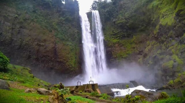 Majestic twin waterfall cascading from a high rocky cliff into a river with a small concrete dam infrastructure.