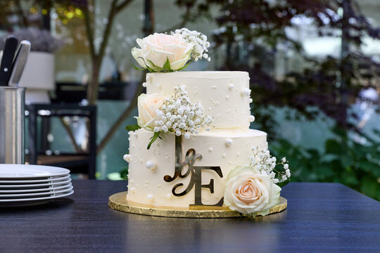 A beautiful two-tier white wedding cake decorated with fresh roses, gypsophila flowers, pearls, and a gold monogram topper on a table.