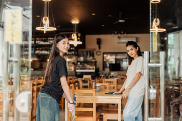 Obraz premium Two cheerful young Asian women trainees perform restaurant opening duties while one smiles at the camera.