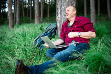 Obraz premium An elderly man with a laptop and a camera sits on the grass leaning against a tree and excitedly rejoices at the good news about the photos he sent online