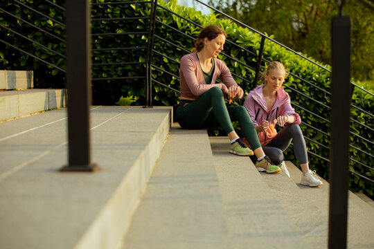Two women rest on steps after workout at outdoor park during sunny day