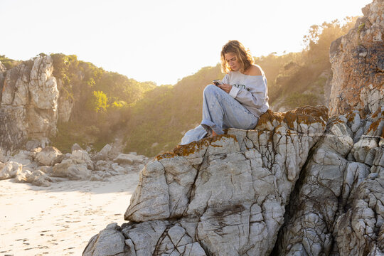 young woman looking at mobile phone sitting on cliff rock at the beach
