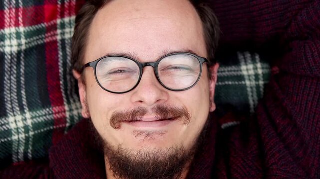Man smiling in glasses lying on plaid blanket close up. Cozy portrait of happy young guy with beard laughing on checkered red picnic blanket, winter comfort and mental health concept. missing tooth