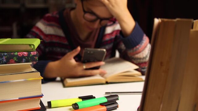 Student using smartphone instead of studying close up. Female student in glasses text message on phone over open book with stacks of textbooks on table, education procrastination and exam preparation