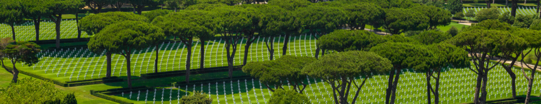 Aerial view of a military cemetery with neat rows of white headstones on green lawns, shaded by large green stone pine trees under bright sunlight. It is a summer sunny morning.
