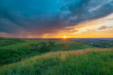 Fototapeta premium Stormy clouds over the horizon. Summer sunset over the green grass field. Approaching rain in the evening. 