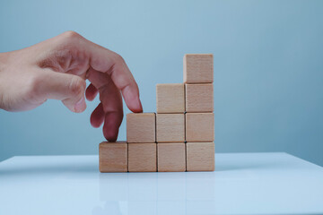 Hand arranging wooden blocks into an upward staircase on a blue background, symbolizing business growth, progress, strategy, career development, achievement, and step-by-step success. © Chaithawat