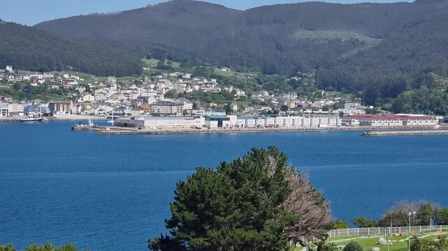 A sweeping panoramic view of Viveiro town curved around the sandy bay and blue sea of the Covas beach with rolling green mountains under a clear sky in Galicia, Spain.