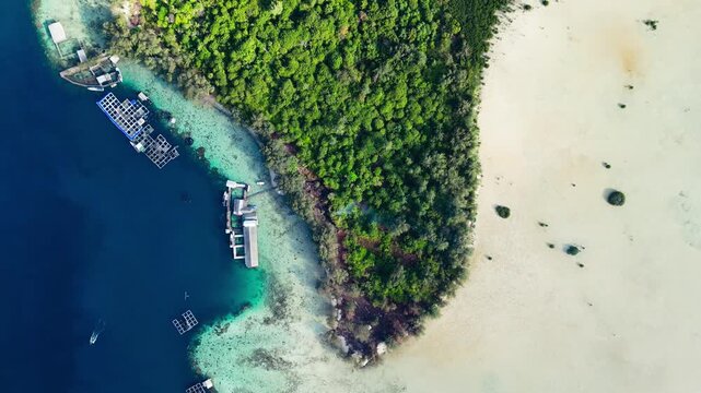 Top-down aerial view of a tropical island featuring a lush green forest bordered by a wide white sandflat on one side and a turquoise coral reef with aquaculture cages on the other