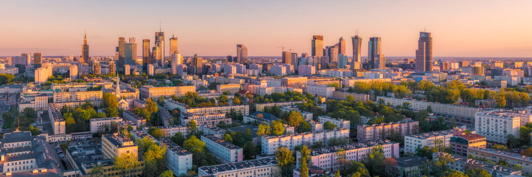 Warsaw, Poland - 20 April 2025: Aerial view of the modern city skyline with skyscrapers, residential blocks, and lush green trees during a golden sunset.