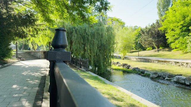 A metal railing guides a stone path next to a calm river in Simferopol, Crimea. This sunlit park scene fills the air with a peaceful and happy feeling.
