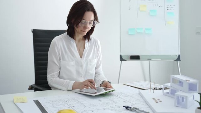 Female architect analyzing blueprints and architectural models in office, using digital tablet for modern design planning and engineering project development