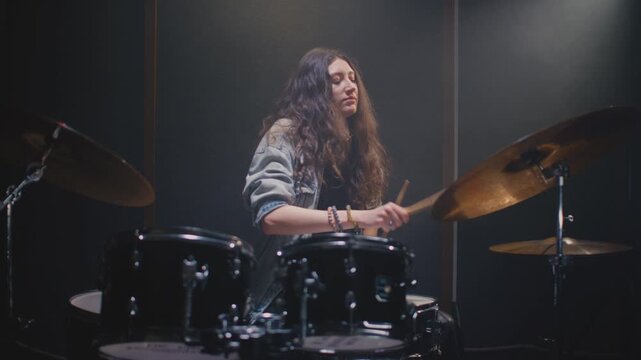 Young drummer girl counts down rhythm with drumsticks, and song starts. Female musician with long curly hair plays a drum kit. Rock band on concert, live on rehearsal room. Repetition in slow motion