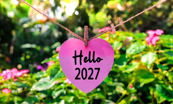 Love pink paper with the text Hello 2027 hanging on a rope in a colorful flower garden. The beginning of 2027. New Year's concept 2027