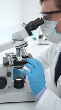 Male masked scientist wearing blue protective gloves, is using microscope doing scientific research in a laboratory with test tubes containing blue liquid. Medicine, healthcare and science concept