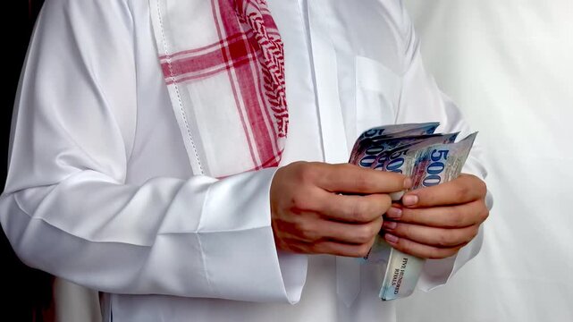 Saudi man wearing traditional thobe counting five hundred riyal banknotes close up.