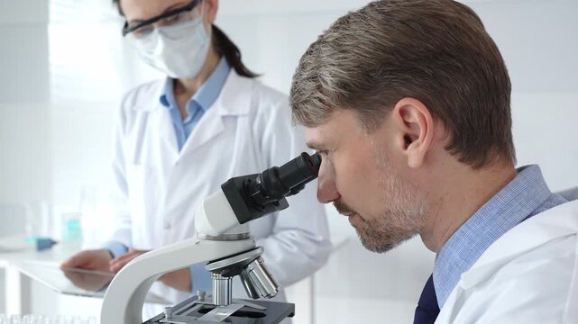Scientific professional examining specimen through microscope, laboratory assistant nearby wearing protective mask, both dressed in white lab coats during research process. Medicine and science