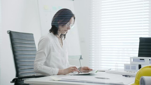 Female architect analyzing blueprints and architectural models in office, using digital tablet for modern design planning and engineering project development