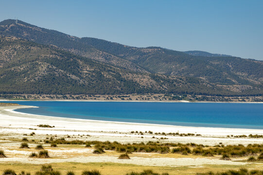 White alkaline beach and turquoise water of Salda lake in morning sunlight. Distant green mountain under clear sky, summer travel destination, Burdur province, Anatolia, Turkey.