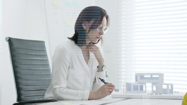 Professional female architect working on blueprints and scale models at a bright office desk, concentrating on urban planning, structural design and project development