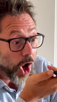 Angry middle-aged businessman with a beard and glasses emotionally arguing and gesturing while recording a voice memo on his mobile phone, showing frustration and stress in a close-up shot