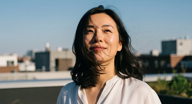 A peaceful Asian woman with a gentle smile looking up towards the sky on an urban rooftop during a sunny day. Her dark hair is slightly tossed by the wind