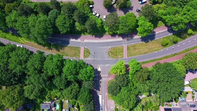 Top down aerial drone hyperlapse footage of a road intersection with cars moving through traffic, bicycle paths and surrounding green trees. Fast motion urban transportation infrastructure.