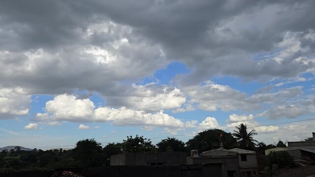 Dark monsoon clouds race across a blue sky, flickering with light. Below, windy treetops & rooftops frame the horizon, perfectly capturing the restless, changing energy of the oncoming stormy weather.