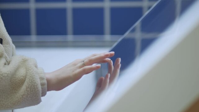 Hands gently touch glass near blue-tiled wall. Soft focus captures quiet, thoughtful moment. Person wears cozy sweater, rings on fingers. Scene evokes calm, introspection, or quiet day indoors