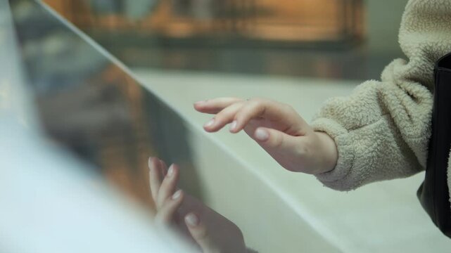 Hands gently touch a reflective glass surface. Soft, cozy sweater visible on arms. Reflections of warm, blurred indoor lights. Calm, curious, and exploratory mood. Suitable for themes like discovery