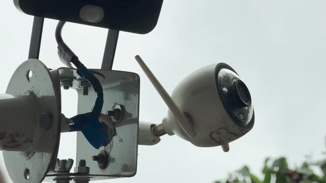 A white security camera records an outdoor area during a rainstorm. Raindrops hit the wet plastic casing. This creates a soft tapping sound.