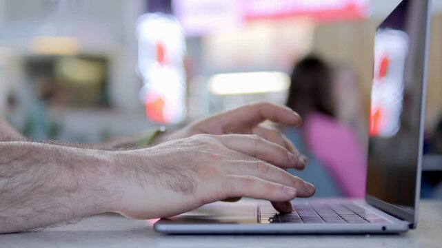 Hands type on laptop keyboard. Background shows blurry cafe or office. Fingers move quickly over keys. Person focused on screen, intent concentration. Could represent remote work, online tasks