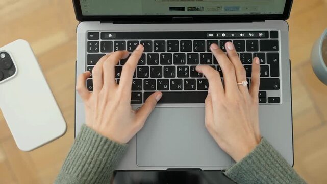 Top view of female hands typing on laptop keyboard next to smartphone on wooden desk Flat lay overhead shot of professional typing on laptop computer with a smartphone