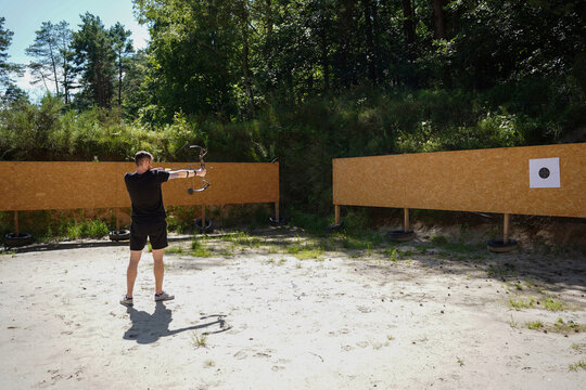Man in black athletic gear aiming a compound bow at a target outdoors on a sunny summer day. Archery sport training, professional bowhunter practice, outdoor shooting range discipline 