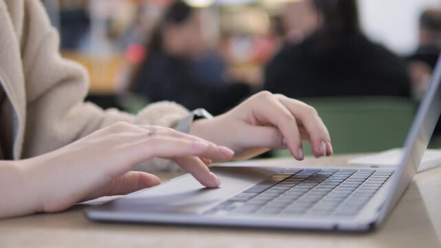 Close-up of hands typing on a laptop keyboard. Fingers move quickly across keys with precision. Background shows blurred people in a public space. Person appears focused and concentrated