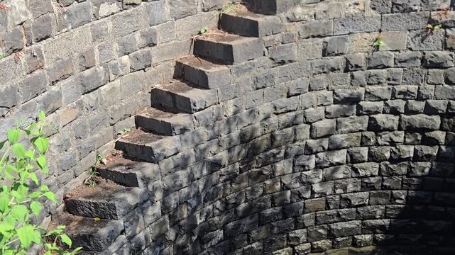 Ancient helical stepwell with spiral stone stairs transitioning into rippling water, showcasing deep shadows, bright sunlight, and a vivid reflection of the architecture's arc and blue sky.