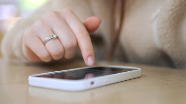 Hand taps smartphone screen. Finger moves across phone. Person wears ring. Phone rests on wooden surface. Focus on interaction with device. Ideal for digital communication, tech usage