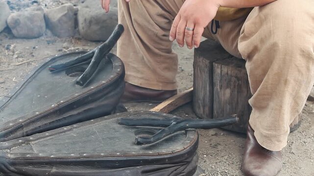 Historical reenactment of an artisan in traditional clothing using antique leather bellows to fan the flames in an ancient outdoor workshop.