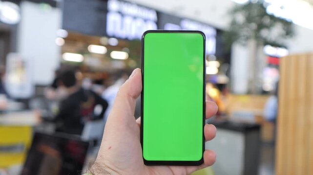 Hand holds phone with green screen in busy indoor space. Background shows blurred people and cafe seating. Ideal for app demos, digital content, or UI mockups