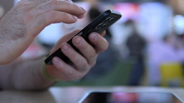 Close-up of hand using smartphone. Finger taps screen actively. Background shows public space blur. Person focused, engaged with device. Ideal for tech, communication, or urban lifestyle themes
