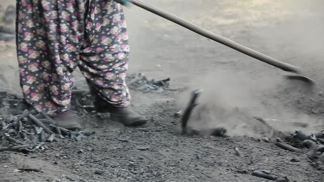 Close-up shot of a person in traditional clothing using a rake to collect hot charcoal in an outdoor production site.
