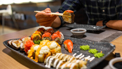 Person holding a sushi roll with chopsticks over a colorful assortment of Japanese sushi and rolls © Valeriia