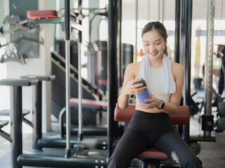 Fototapeta premium Smiling young Asian woman in sportswear holding a water bottle and sitting on a bench press in the gym after a workout.