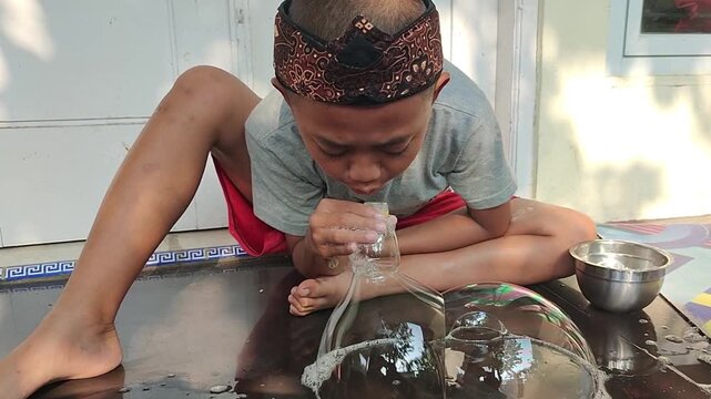 An Indonesian boy wearing a traditional  sundanese batik headband, a casual grey t-shirt and red shorts sits on the floor, playing soap bubble using a plastic bottle  