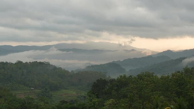 Beautiful sunrise time lapse of foggy tropical mountains, majestic hills covered in moving thick mist under a dramatic cloudy sky in Cilacap, Indonesia.