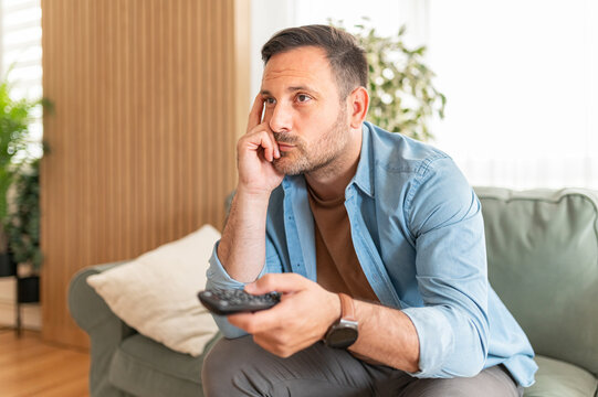 Man feeling boredom and indifference while relaxing on a comfortable piece of furniture, holding a remote control and appearing disengaged with the digital media