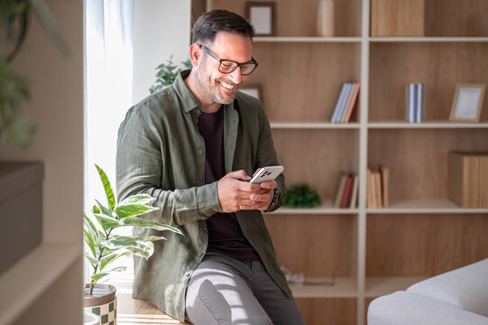 Smiling man relaxing at home, enjoying wireless social media connection, holding a mobile device and actively messaging friends on a communication application