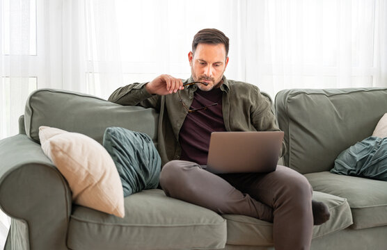 Man sitting on couch, holding eyeglasses, concentrating during remote work, using laptop for online business communication and education at residence