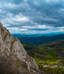 Obraz premium Dramatic mountain view from a rocky cliff overlooking green valleys under a stormy, cloudy sky. Epic landscape of high green mountains and rocky peak before a storm.