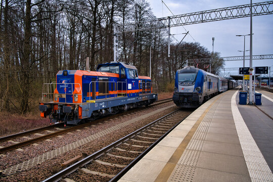 A blue and orange diesel shunting locomotive and an electric passenger train on the platform of the railway station in the seaside town of Świnoujście.